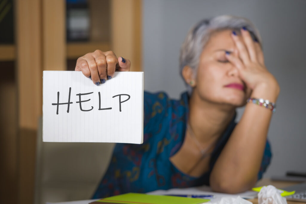 desperate and stressed attractive middle aged Asian woman holding notepad asking for help feeling overworked and exploited working at computer office desk suffering stress and fatigue